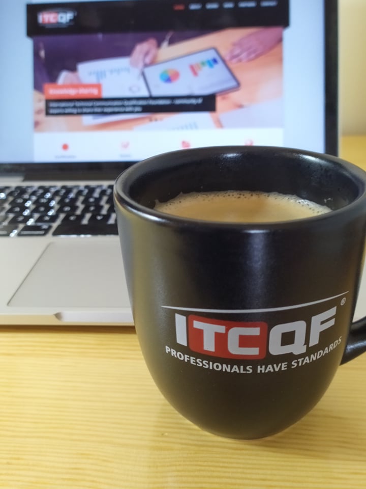Kubek z logo ITCQF na tle laptopa. W kubku jest pyszna kawa z mlekiem.